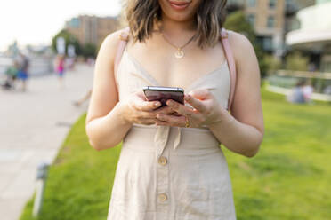 Nahaufnahme einer Frau, die in einem öffentlichen Park steht und mit ihrem Smartphone SMS schreibt - WPEF03403