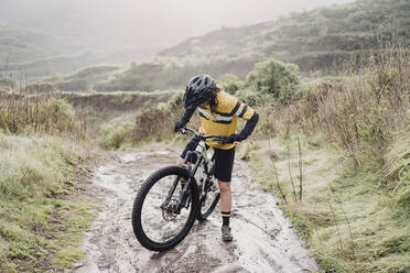 Ganzkörper-Radfahrerin auf schlammigem Bergpfad an einem regnerischen Tag - CAVF89228