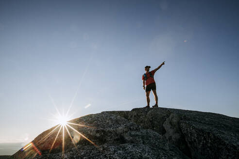 Männlicher Wanderer zeigt auf Aussicht vom felsigen Gipfel eines Berges mit Sonnenstern - CAVF89237