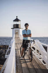 Männlicher Radfahrer posiert mit Fahrrad vor einem Leuchtturm an der Küste von Maine - CAVF89243