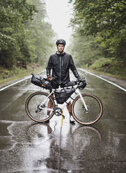 Male bike packer stands with bike in road in pouring rain, Maine - CAVF89244