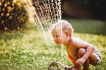 Young boy sticking tongue out drinking water from sprinkler in garden - CAVF89245