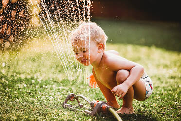 White boy sticking tongue out drinking water from sprinkler in garden - CAVF89246