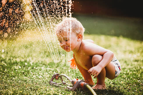Weißer Junge streckt die Zunge heraus und trinkt Wasser aus dem Rasensprenger im Garten - CAVF89246