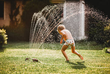 Junge weiße Junge läuft unter dem Wasser aus dem Sprinkler im Garten - CAVF89247