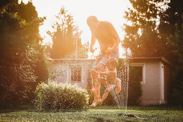 Vater und Kind springen über das Wasser aus dem Rasensprenger im Garten - CAVF89248