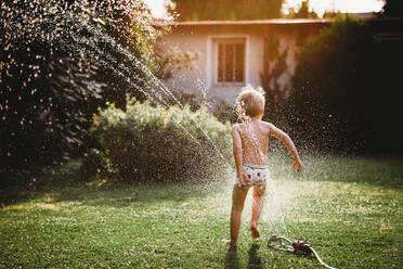 Young white boy running under the water from the sprinkler in garden - CAVF89249