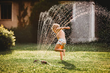 Young white boy running under the water from the sprinkler in garden - CAVF89250