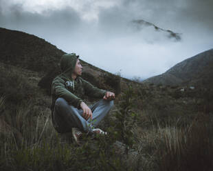 Junger Mann sitzt auf einem Felsen und bewundert Berge mit tief hängenden Wolken - CAVF89268