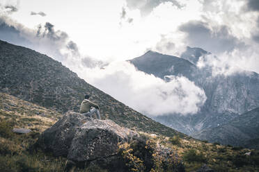 Junger Mann sitzt auf einem Felsen und bewundert Berge mit tief hängenden Wolken - CAVF89269