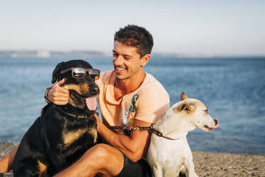 Lächelnder erwachsener Mann, der mit Hunden spielt, während er am Strand gegen den Himmel sitzt - DCRF00872