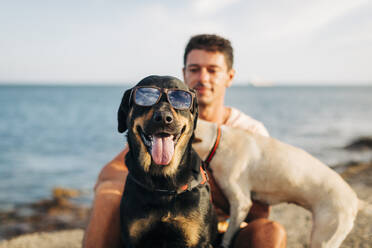 Hund mit Sonnenbrille streckt die Zunge heraus, während er mit einem Mann am Strand sitzt - DCRF00873