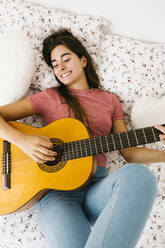Junge Frau liegt auf dem Rücken und spielt Gitarre im Schlafzimmer - XLGF00545