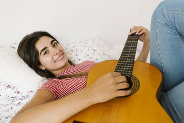 Young woman lying down while playing guitar in bedroom - XLGF00546