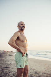 Man with hand on hip standing at beach during sunset - MEUF02085