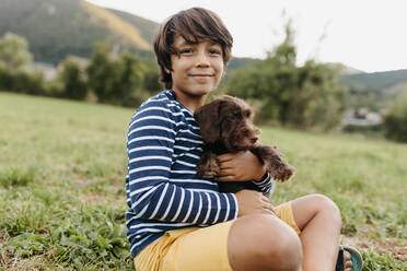 Boy holding puppy while sitting on grass at backyard - VABF03505