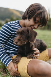 Smiling boy playing with puppy while sitting on grass at backyard - VABF03507