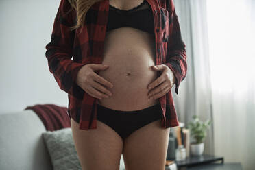 Nahaufnahme einer schwangeren Frau mit Händen auf dem Bauch, die zu Hause steht - BZF00574