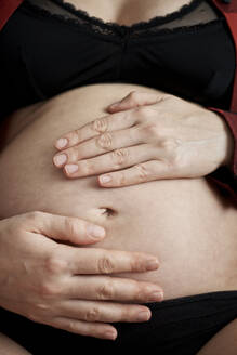 Nahaufnahme einer schwangeren Frau mit Händen auf dem Bauch, die zu Hause sitzt - BZF00577