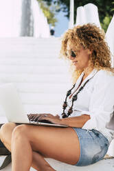 Frau mit lockigem Haar und Sonnenbrille, die einen Laptop benutzt, während sie auf einer Treppe sitzt - SIPF02184