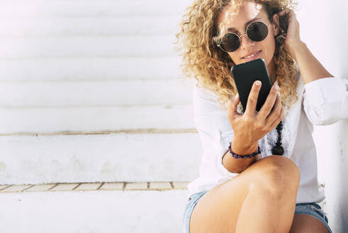 Schöne Frau mit Sonnenbrille, die ein Smartphone benutzt, während sie auf einer Treppe sitzt - SIPF02186