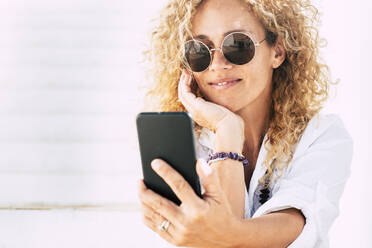 Nahaufnahme einer Frau mit Sonnenbrille, die ein Smartphone benutzt, während sie im Freien sitzt - SIPF02188