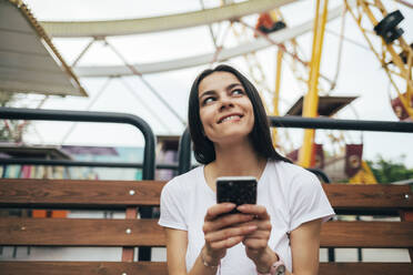 Junge Frau benutzt ihr Smartphone, während sie auf einer Bank im Vergnügungspark sitzt - OYF00202