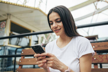 Nahaufnahme einer lächelnden Frau, die ein Mobiltelefon benutzt, während sie auf einer Bank im Vergnügungspark sitzt - OYF00203