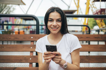 Schöne Frau mit Smartphone auf einer Bank im Vergnügungspark sitzend - OYF00204