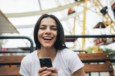 Frau hält Smartphone lachend auf einer Bank im Vergnügungspark sitzend - OYF00205