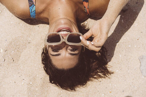 Cheerful woman wearing sunglasses while lying at Valdevaqueros beach on sunny day, Tarifa, Spain - DAMF00465