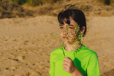 Thoughtful girl holding flowers while standing at beach during sunny day - ERRF04412