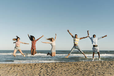 Cheerful young friends jumping with hand raised on beach during sunny day - RDGF00154