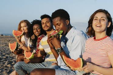 Freunde genießen ein Stück Wassermelone, während sie an einem sonnigen Tag am Strand sitzen - RDGF00169