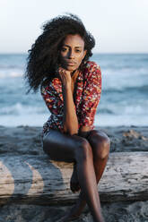 Junge Frau mit Hand am Kinn am Strand sitzend an einem sonnigen Tag - RDGF00188
