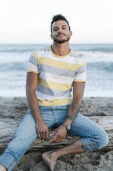 Junger Mann sitzt auf einem Baumstamm am Strand - RDGF00194