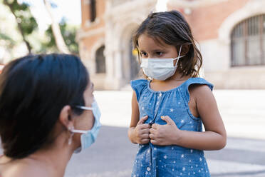 Girl wearing mask looking at mother while standing on street - EGAF00740