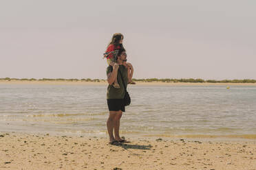 Vater, der seine Tochter auf den Schultern trägt, während er am Strand steht, gegen einen klaren Himmel an einem sonnigen Tag - ERRF04423