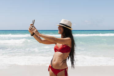 Woman wearing bikini taking selfie with smart phone while standing against sea - EGAF00754