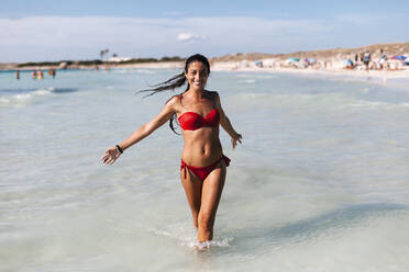 Cheerful mid adult woman wearing bikini walking in sea against sky - EGAF00758
