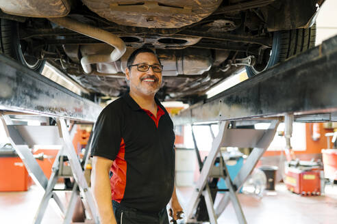 Smiling male mechanic standing below car in auto repair shop - EGAF00772