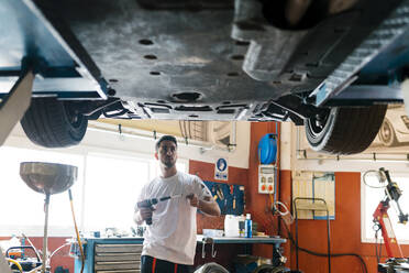 Young mechanic holding work tool looking at car while standing in auto repair shop - EGAF00780