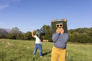 Boys playing with robot cardboard box while standing in meadow - VABF03525