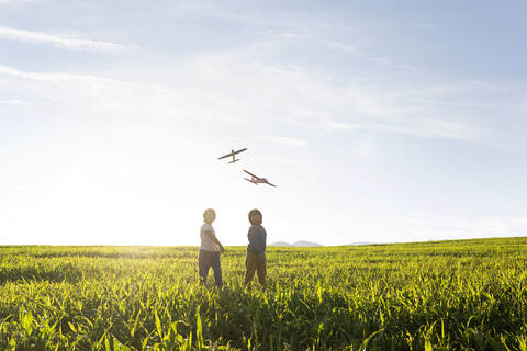 Boys looking at flying airplane toy while standing on grass in meadow stock photo