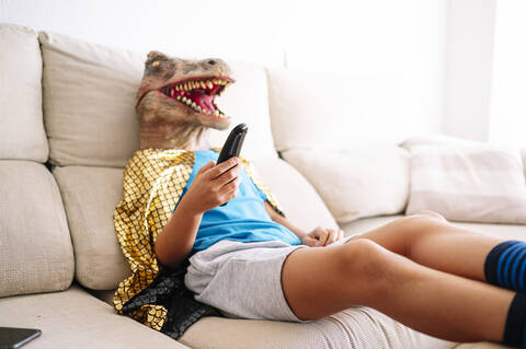 Junge mit Dinosauriermaske sieht fern, während er sich zu Hause auf dem Sofa entspannt, lizenzfreies Stockfoto