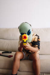 Boy wearing alien mask playing with toy gun while sitting on sofa in living room - JCMF01456
