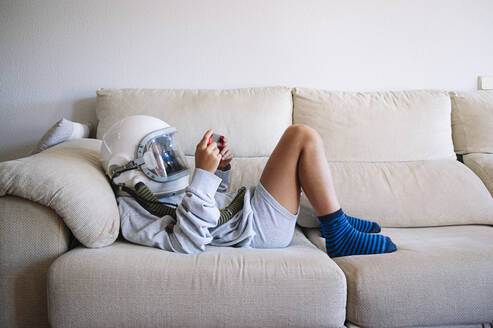 Boy wearing space helmet playing video game while lying on sofa at home - JCMF01458