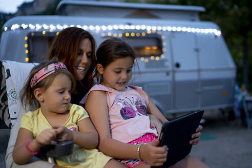 Lächelnde Mutter mit Töchtern, die ein digitales Tablet benutzen, während sie in der Abenddämmerung an einem Wohnmobil sitzen - OCMF01725