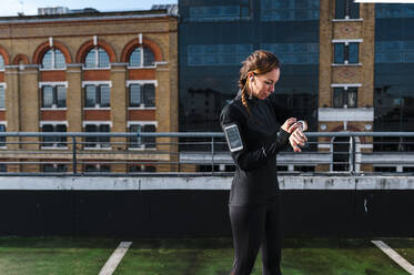 Female athlete checking smart watch while standing on rooftop in city - JMPF00405