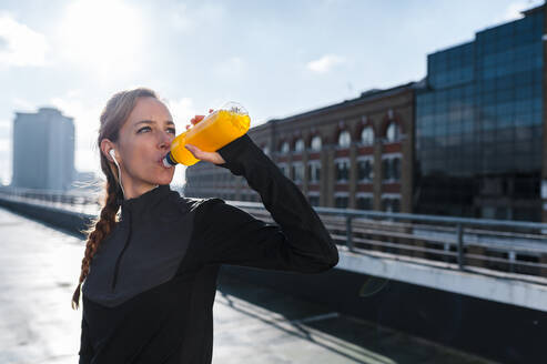 Female athlete having drink while standing on rooftop against sky during sunny day - JMPF00413